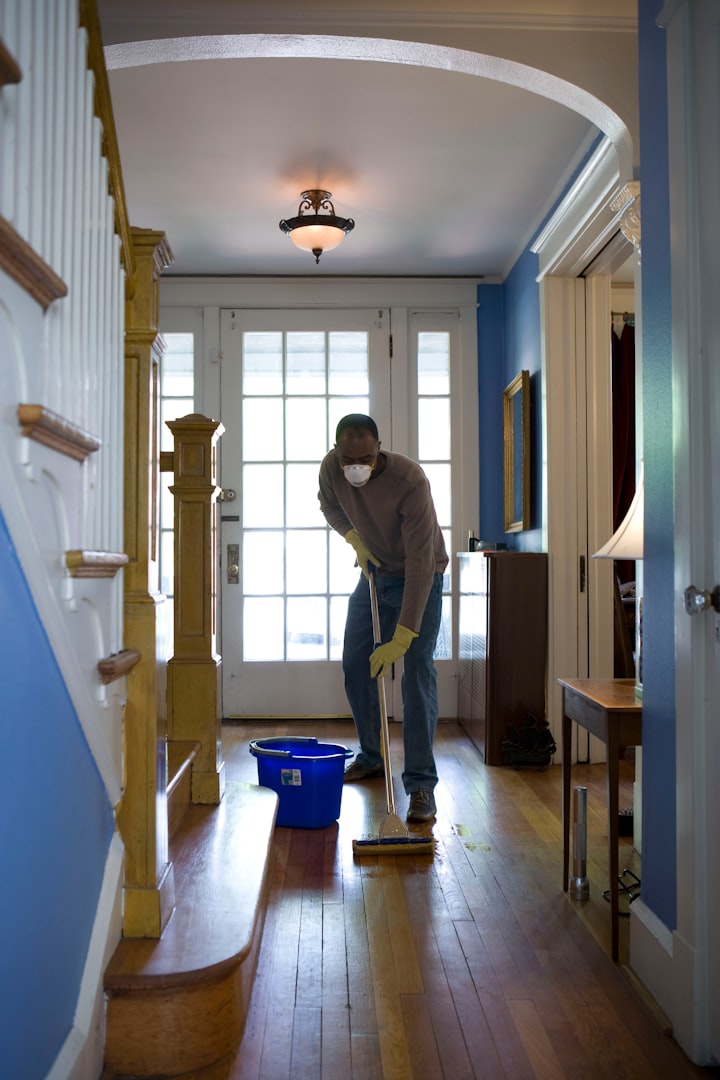 A woman wearing yellow cleaning gloves is wiping down a wooden desk in a bright, modern office space with large windows and plants in the background. She is focused on her task, and the room is well-lit by natural light.